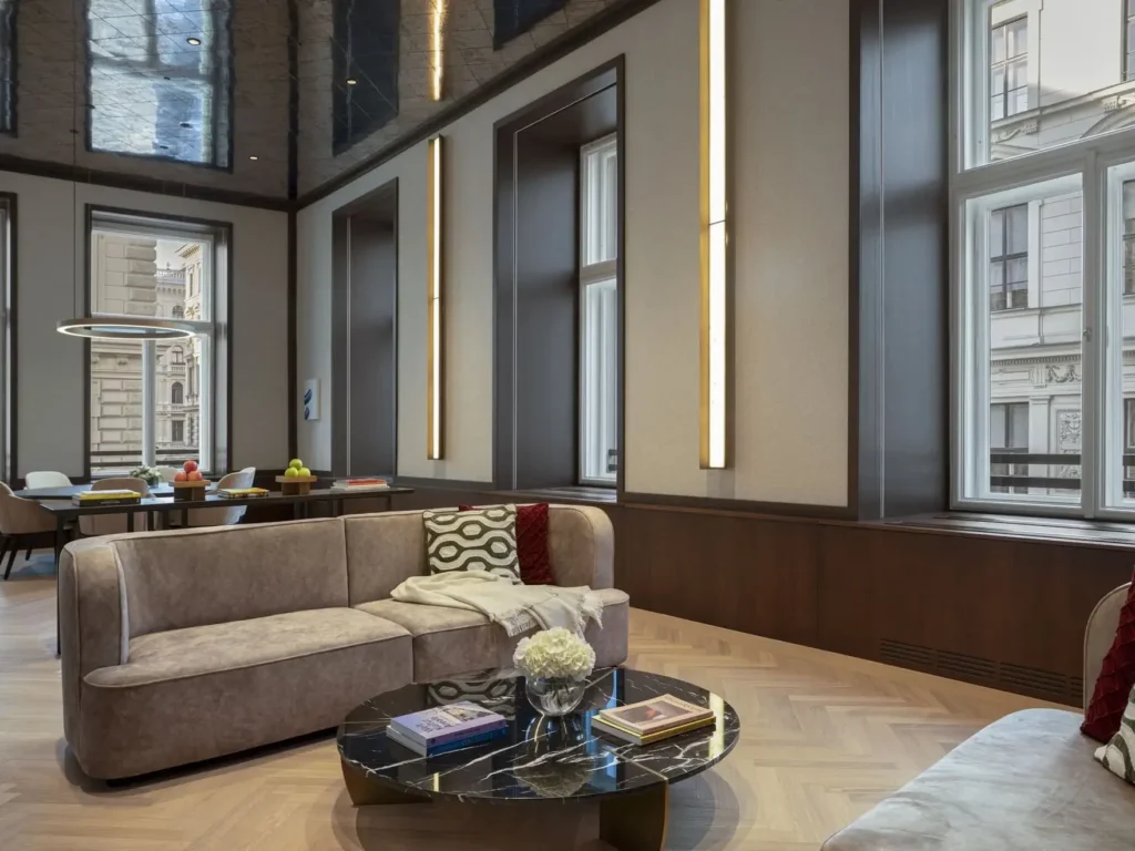 Moderner Lounge-Bereich mit beigen Sofas, einem schwarzen Marmor-Couchtisch, Büchern und großen Fenstern; kunstvoll gestaltet im Stil einer Wiener Suite, mit einem Essbereich im Hintergrund.